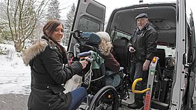 A woman and a man help a person in a wheelchair into a van.