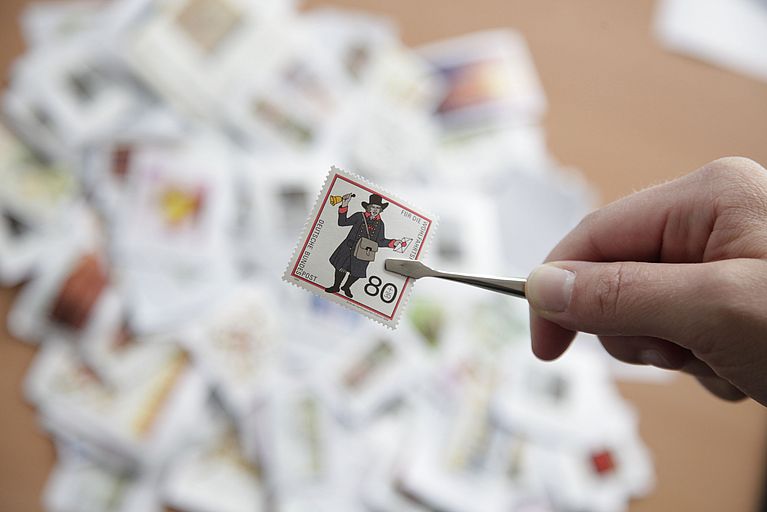 Eine Briefmarke wird mit einer Pinzette in die Kamera gehalten. Im Hintergrund liegen viele weitere Briefmarken auf einem Tisch.