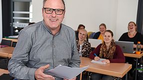 Teacher stands in front of the students and smiles at the camera.