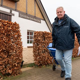 Andreas Zylla geht auf dem Bürgersteig an einem Haus vorbei. Er zieht einen Rollwagen hinter sich her. In der linken Hand hält er eine Zeitschrift.