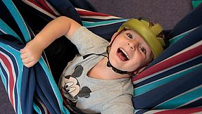 Kind mit Epilepsiehelm liegt in einer Schaukel und lacht.