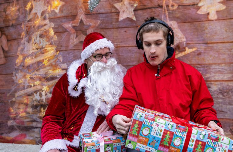Der Nikolaus schaut gespannt zu, wie ein junger Mann ein Geschenk auspackt. Beide sitzen auf einer Bank. Im Hintergrund ist Weihnachtsdeko.