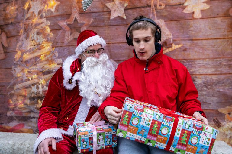 Der Nikolaus schaut gespannt zu, wie ein junger Mann ein Geschenk auspackt. Beide sitzen auf einer Bank. Im Hintergrund ist Weihnachtsdeko.
