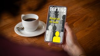 Eine Hand hält ein Smarthphone mit der digitalen RING-Ausgabe. Daneben steht eine Tasse Kaffee.