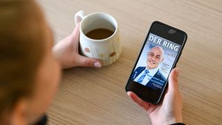 Eine Person sitzt an einem Tisch und hält ein Smartphone mit der digitalen Ausgabe vom RING in der Hand. Auf dem Tisch steht eine Tasse mit Kaffee.
