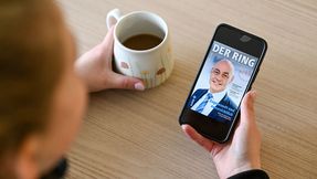 Eine Person sitzt an einem Tisch mit einer Tasse Kaffee und hält ein Smartphone mit der digitalen Ausgabe von DER RING in der Hand.