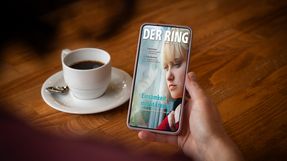 Eine Person sitzt an einem Tisch mit einer Tasse Kaffee und hält ein Smartphone mit der digitalen Ausgabe von DER RING in der Hand.
