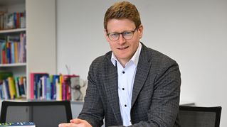 Dr. Simon Stark sitzt an einem Tisch und lächelt in die Kamera.