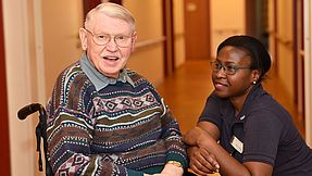 Eine Pflegekraft lehnt sich an den Rollstuhl eines Senioren und lächelt ihn an.