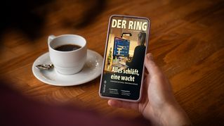 Jemand sitzt an einem Tisch mit einer Tasse Kaffee und hält ein Smartphone mit der neuen Ausgabe von DER RING in der Hand.
