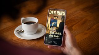Jemand sitzt an einem Tisch mit einer Tasse Kaffee und hält ein Smartphone mit der neuen Ausgabe von DER RING in der Hand.