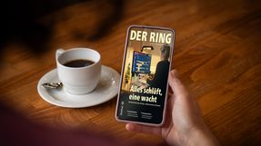 A person is sitting at a table holding a smartphone with the digital version of DER RING in her hand.